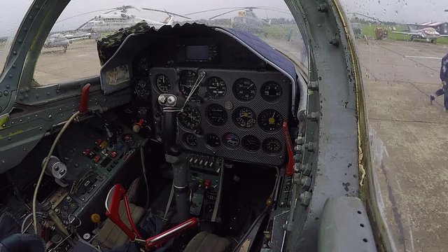 Cabin of combat fighter plane. Inside view of cockpit and dashboard close up. The background of different helicopters at the old airfield. Some people walk around.