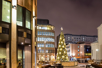 Christmas tree is decorated with lights, balls, toys and garlands, and star of gold color at the top. Facades of modern buildings in the background. Winter evening.