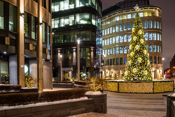 Christmas tree is decorated with lights, balls, toys and garlands, and star of gold color at the top. Facades of modern buildings in the background. Winter evening.