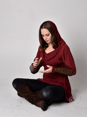 full length portrait of a brunette girl wearing a red fantasy tunic with hood. Sitting pose on a...