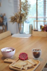 Three Slice breads with half dragon fruit in a wooden plate , a yogurt bowl on the left side and a glass of ice americana is on the right side (spot selective focus)