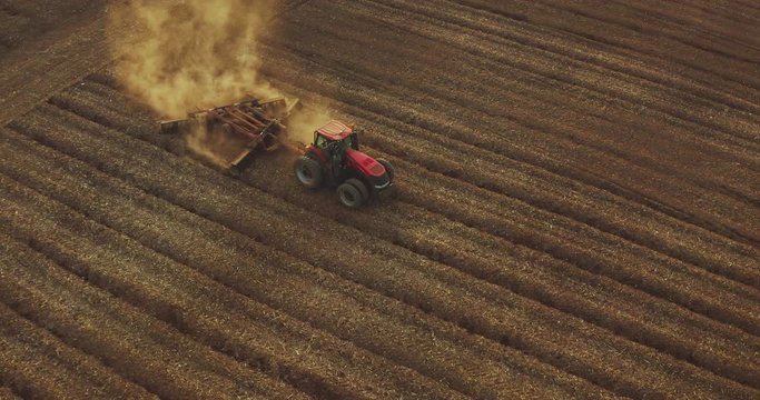 Aerial view of a farmer driving a red tractor through dusty fields at sunset with golden dirt rising into the air, farming at sunset, climate change barren dusty fields