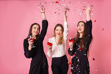 Three women celebrate the New Year party having fun laughing under the flying confetti and drinking...