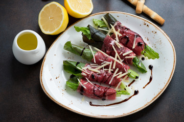 Carpaccio made of raw marbled beef with salad leaves and grated parmesan, studio shot on a dark brown metal surface