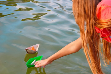 A girl launches colored paper boats in the water at sunset. Cardboard ships in a fountain in a city park. Children's outdoor recreation in the summer.