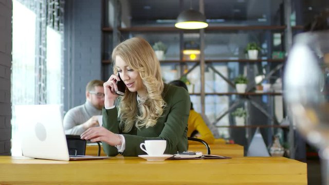 Tracking shot of smiling blonde businesswoman speaking on mobile phone and using laptop computer at cafe table on lunch