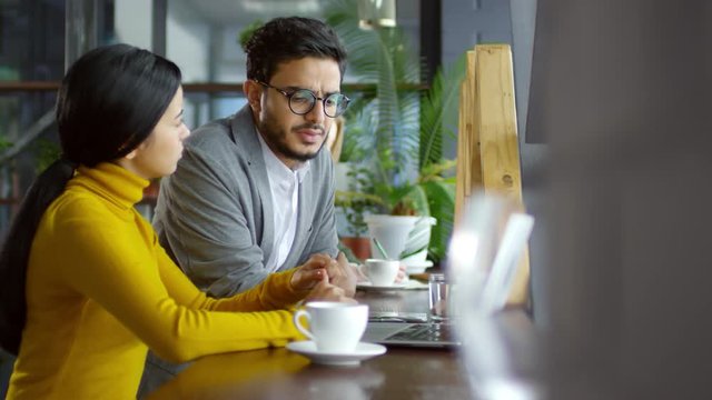 Middle eastern businessman speaking with female Asian colleague while sitting together at cafe table beside the window and working on laptop