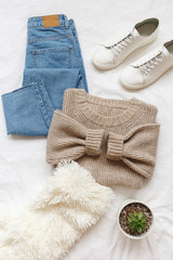 Blue jeans, beige knitted sweater, sneakers, fur scarf, home plant in a pot lying on bed on white sheet. Overhead view of simple basic woman's casual outfit. Trendy women clothes. Flat lay, top view.