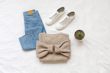 Blue jeans, beige knitted sweater, sneakers, home plant in a pot lying on bed on white sheet. Overhead view of simple basic woman's casual day outfit. Trendy stylish women clothes. Flat lay, top view.