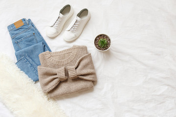 Blue jeans, beige knitted sweater, sneakers, fur scarf, home plant in a pot lying on bed on white sheet. Overhead view of simple basic woman's casual outfit. Trendy women clothes. Flat lay, top view.
