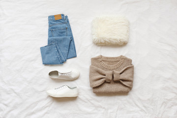 Blue jeans, beige knitted sweater, sneakers, fur scarf lying on bed on white sheet. Overhead view of simple basic woman's casual day outfit. Trendy women clothes. Hipster look. Flat lay, top view.