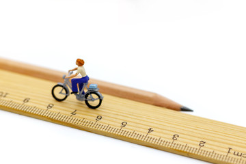 Miniature people ride bicycles on wooden ruler.