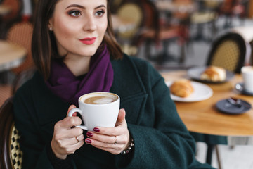 Beautiful young brunette woman with short hair wearing green coat and purple scarf sitting at a table in cozy street outdoor cafe and drinking coffee. Girl with cup of cappuccino at restaurant terrace