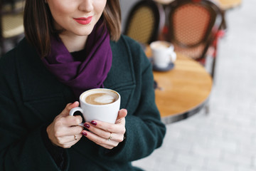 Cup of coffee in women's hands. Beautiful young brunette woman wearing green coat sitting at a table in cozy street outdoor cafe and drinking coffee. Girl with cup of cappuccino at restaurant terrace.