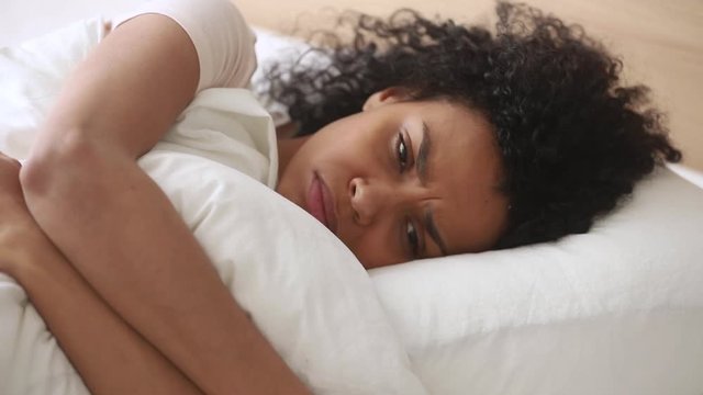 Depressed african woman lying on bed embraces pillow feels unhappy