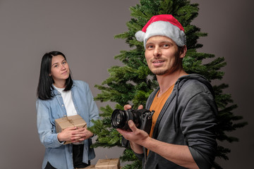 A young guy in a Christmas cap is filming on a mirrorless camera as a young girl with black hair is wrapping Christmas presents. 