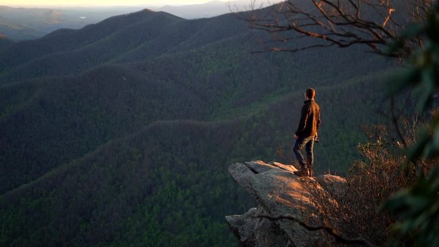 Hiking man is backlit by the setting sun as he stands on a cliff and looks out over the mountains and forest below