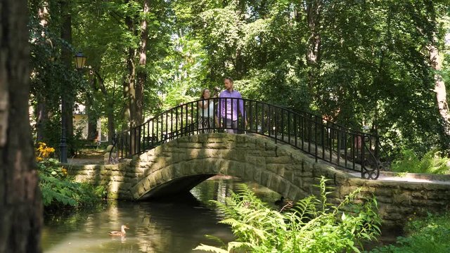A young couple walks trough small stone bridge in park, she points something in front, he noticed other thing and they stop, she is excited, they kiss each other and walk away