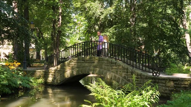 A young couple walks trough small stone bridge in park, stops in the middle and starts talking cheerfully, she is expressive and smiling. They walk away after a while