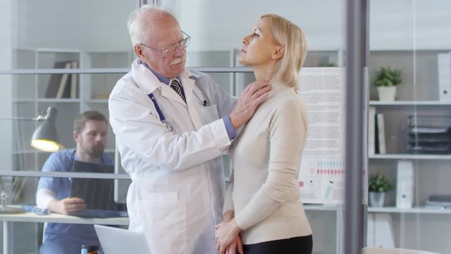 Senior doctor in white coat examining neck and throat of female patient and speaking with her during medical checkup in clinic; male physician working with x-ray image in background