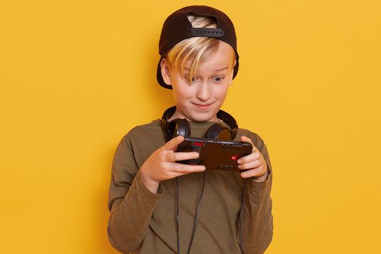 Portrait of emotional and active little boy with blond hair and green shirt, carrying his finger on screen of smartphone while playing his favorite online game, poses over yellow background in studio.