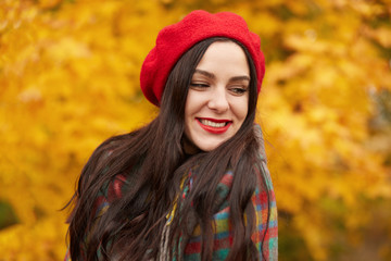 Close up portrait of young fashionable girl wears red beret, having dark long straight hair, resting in park in autumn stands around yellow trees and smiling. Trendy young woman in fall in fall forest