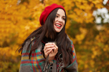 Happy woman wearing stylish red beret and wrapped warm checkered blanket, looking aside and laughing, holding thermos cup of coffee or tea, spending time in open air, enjoying autumn days in park.