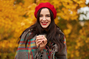 Fashion woman walking in autumn park wrapped in checkered warm blanket and red beret, holding thermos cup in hands and drinking hot beverage, enjoying beautiful nature and fall days. Wekend concept.