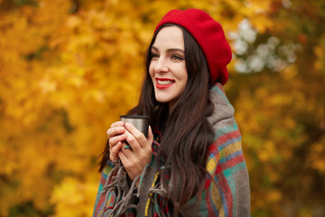 Fototapeta na wymiar Cold season, hot drinks and people concept. Outdoor portrait of beautiful happy young woman wearing red beret and wrapped in blanket drinking coffee or tea from thermos cup, standing in autumn park.