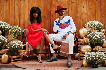 Stylish trendy afro france couple posed together at autumn day. Black african models in love sitting against wooden decoration with flowers and pumpkins.