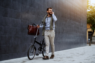 Smiling cheerful caucasian fashionable businessman listening music and pushing his bicycle. In background is gray wall.