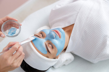 Beautician applying blue clay mask on lady face