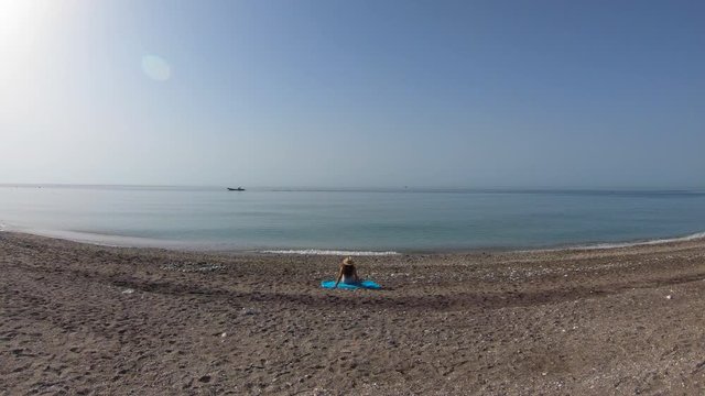 Aerial shot of a young woman in a swimsuit with a hat sitting on the shore of the beach.