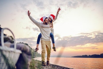 Playful caucasian couple having piggyback on coast next to river. Both having santa hats on heads. In foreground is picnic basket. In background is sunset.