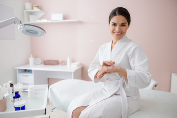 Cheerful young woman in bathrobe applying hand cream