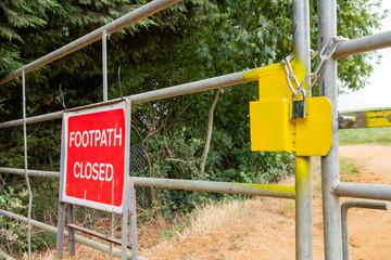 Shallow focus of a yellow painted lock system seen attached to a gate at the entrance to a public footpath. A Closed sign is attached due to a nearby landslip.