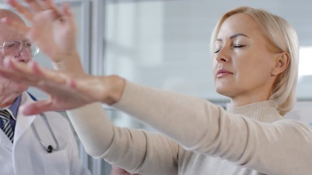 Middle aged caucasian woman standing with eyes closed, touching nose with finger and then clenching fists while senior doctor speaking and looking at her during proprioception test