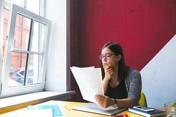 Serious successful business woman in stylish glasses reading paper documents while sitting at desktop in office interior. Female executive director reading resume during work day in company