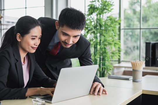 Confidence Young Asian Business Man In Suit Leaning Over A Businesswoman Typing On Laptop Working Together At A Bright Office Desk.