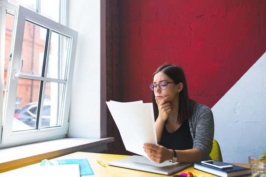 Serious successful business woman in stylish glasses reading paper documents while sitting at desktop in office interior. Female executive director reading resume during work day in company