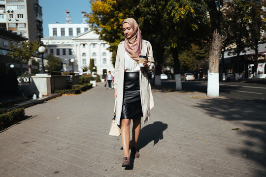 muslim woman in european skirt walks with coffee and bag on a city street