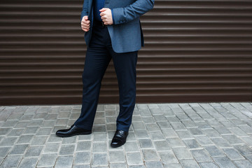 Man's legs. Man wearing suit jacket, pants, blue shirt and black shoes standing near brown roller door on the city street. Details of classic elegant formal look. Trendy men's outfit. Street fashion.