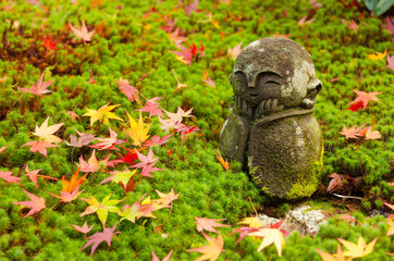 traditional smiling little stone or Jizo buddha monk statue with colorful red maple leaves on green grass ground in Japanese garden with during sunrise , autumn season at Enkoji temple in Kyoto, Japan