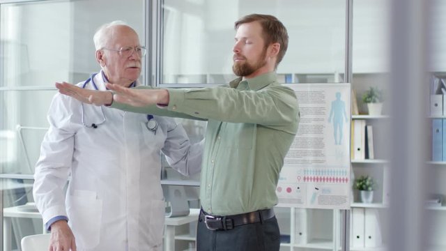 Young caucasian man standing with arms raised and eyes closed and then touching nose and clenching fists while senior neurologist examining him during medical checkup in clinic