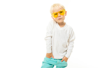 Little boy with short blond hair, blue eyes, cute appearance, in white jacket, light blue pants, stands with yellow glasses and poses beautifully on camera