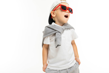 A little boy with short blonde hair, blue eyes, cute appearance, in a white coat around his neck, grey pants, grey cap, glasses from a game with a red frame and black stacks, stands and poses