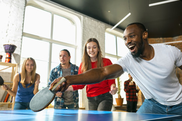 Young people playing table tennis in workplace, having fun. Friends in casual clothes play ping pong together at sunny day. Concept of leisure activity, sport, friendship, teambuilding, teamwork. - obrazy, fototapety, plakaty