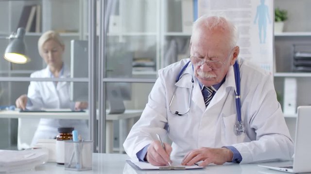 Portrait of cheerful senior doctor in eyeglasses and white coat taking notes at desk and then looking at camera and smiling while working in clinic