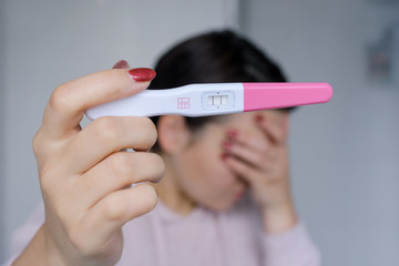 Sad woman holds a positive pregnancy test and covers her face with her hand. Unwonted pregnancy.