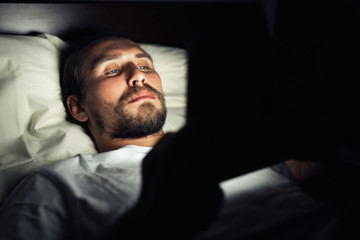 Young handsome and tired man with a beard cannot sleep and is watching something on his tablet at night.